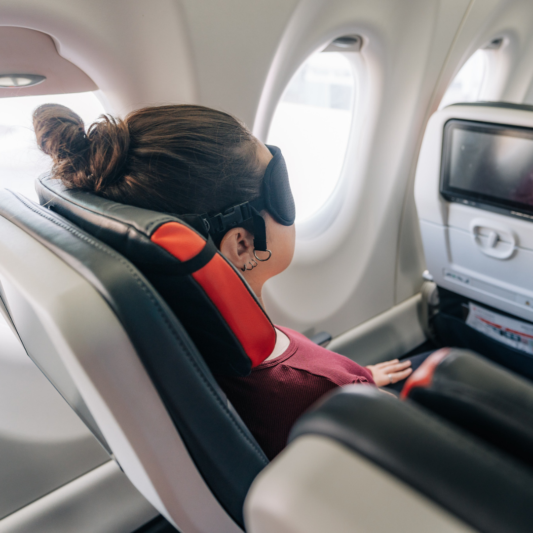 Side view of Woman sleeping while sitting in a plane seat with ALLasleep travel accessory installed on the headrest and on her face.