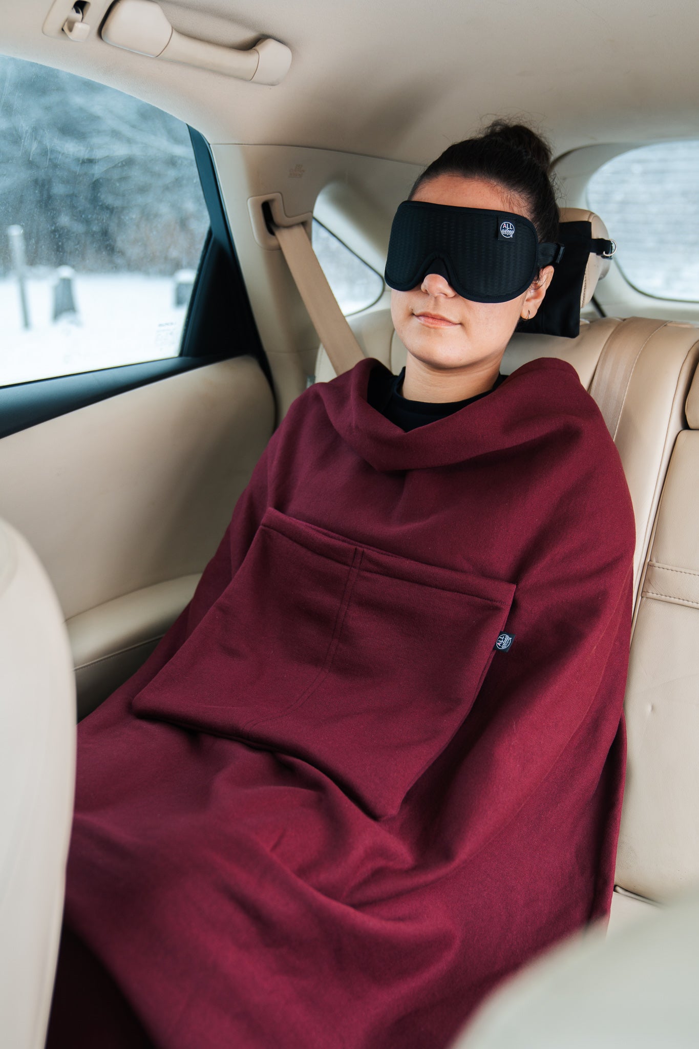 Full view of a woman sleeping in a car wearing her travel blanket and ALLasleep travel accessory